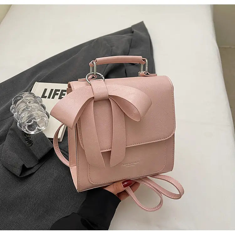 Zartrosa Umhängetasche aus Kunstleder mit Schleifendetail und silberfarbenen Beschlägen, gestaltet als modische Handtasche.
