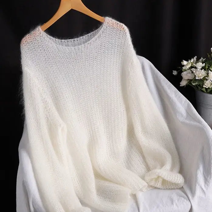 Ein weiches off-weißes Pulloveroberteil in offener Strickoptik, als Blusenstil gestricktes Oberteil, aus Mohair, mit lockerer, fließender Silhouette, hängt auf einem Holzkleiderbügel.