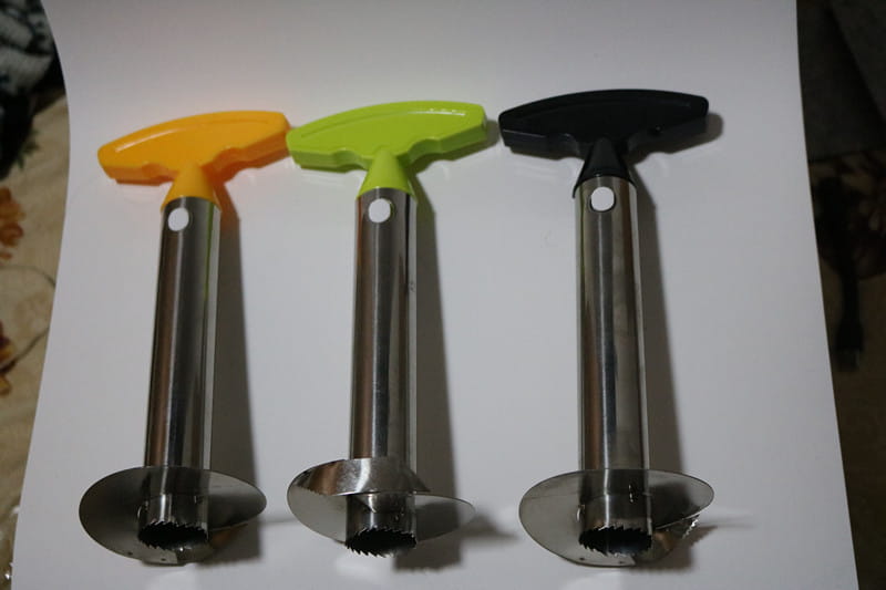 Tres peladores, descorazonadores y herramientas de corte para piña de acero inoxidable con hojas dentadas y mangos con forma de T de colores en naranja, verde lima y negro.