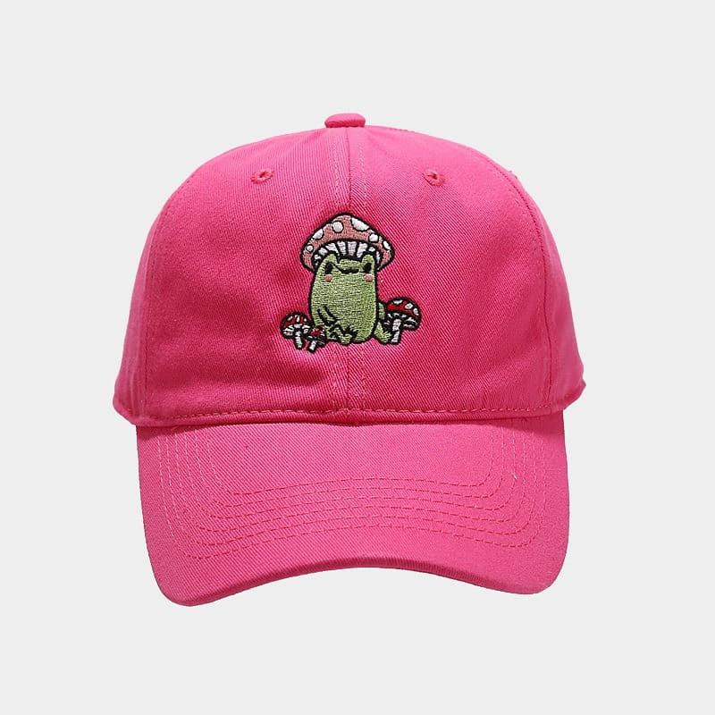 A vibrant pink baseball cap featuring a charming embroidered design of a green frog sitting under a red-capped mushroom with small mushrooms around it.