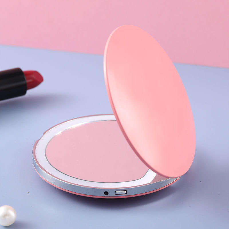 Espejo de maquillaje LED portátil en color rosa, compacto y plegable con un borde metálico plateado y un puerto de carga USB.