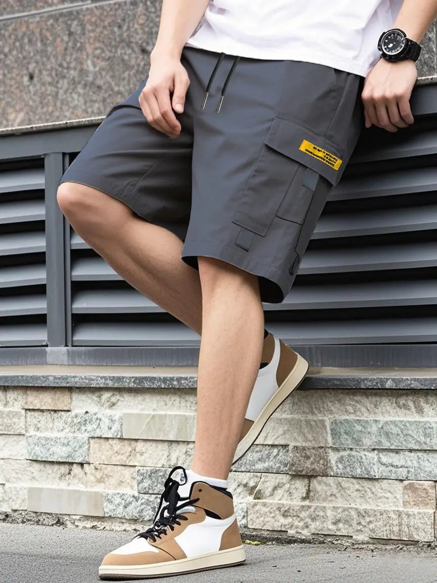 Shorts cargo utilitaires pour homme gris foncé, avec une étiquette jaune sur la poche latérale, présentant un design décontracté d’été, avec cordon de serrage et plusieurs poches.