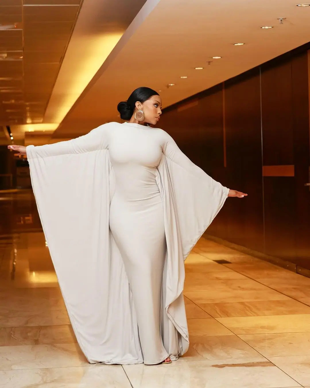 An off-white satin asymmetrical batwing sleeve maxi dress with wide dramatic sleeves and a form-fitting silhouette.