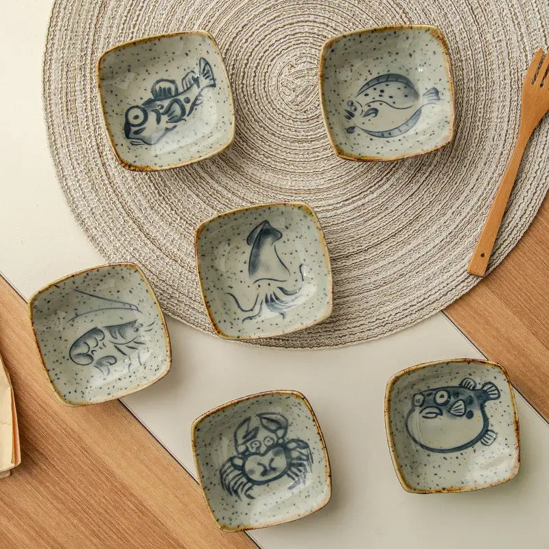 Set of five Japanese style ceramic sea creature sauce dishes featuring unique blue hand-painted designs on speckled off-white glaze with rustic brown rims.