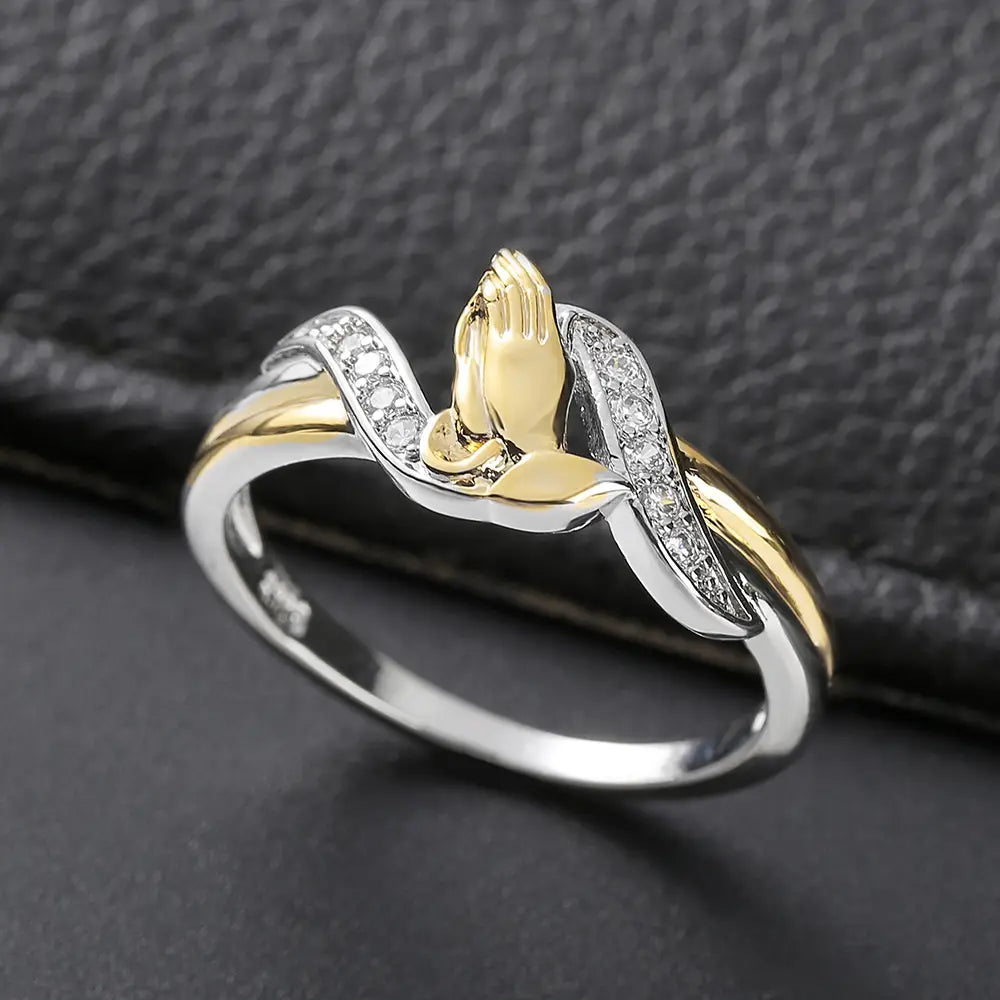 A delicate two tone 18k gold ring with a central praying hands motif set between intertwined silver bands adorned with clear cubic zirconia stones.