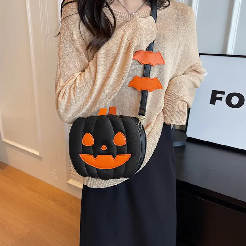 Black pumpkin-shaped crossbody bag with orange stitched facial features and bat appliqués on the strap featuring a detachable charm.
