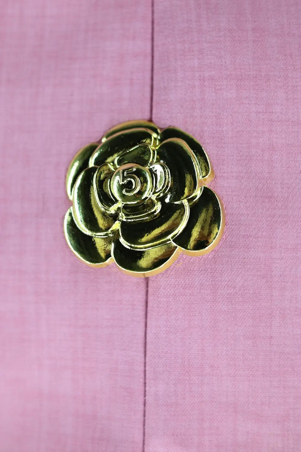 Un bouton brillant de couleur or, en forme de camélia, à pétales superposés et avec un numéro 5 en relief, sur une robe mini sans manches et dos nu, avec des boutons décoratifs en forme de rose dorée