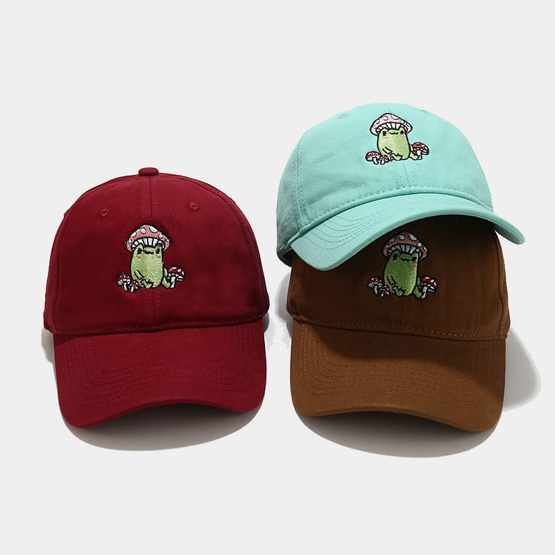 Two embroidered baseball caps in maroon and light blue with brown brim featuring a cute green frog wearing a pink mushroom hat in the frog mushroom embroidered baseball cap collection.