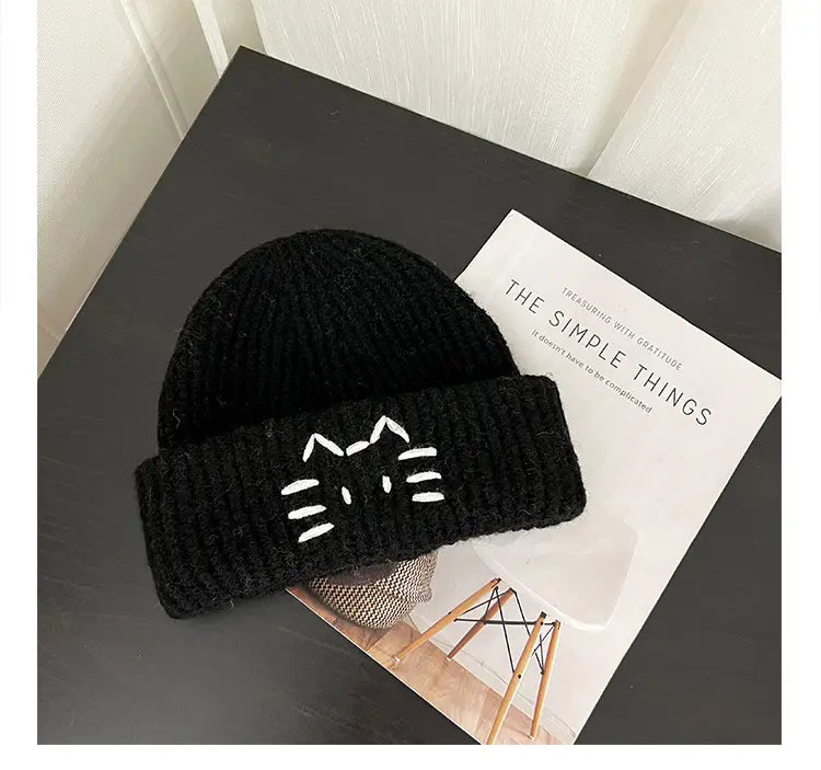 Black knitted beanie hat featuring a white embroidered cat face on the folded brim.