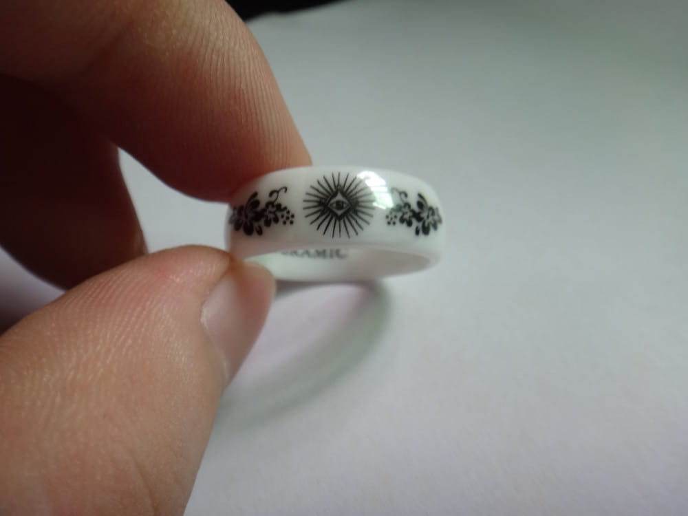 White ceramic ring adorned with black floral and sunburst patterns shown alongside a black tungsten wedding ring with green aluminum 8mm band.