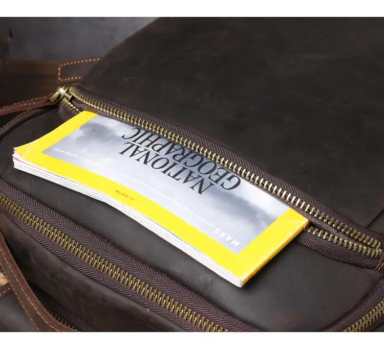 Men’s vintage leather backpack in dark brown cowhide with brass zipper and visible stitching containing a national geographic magazine.