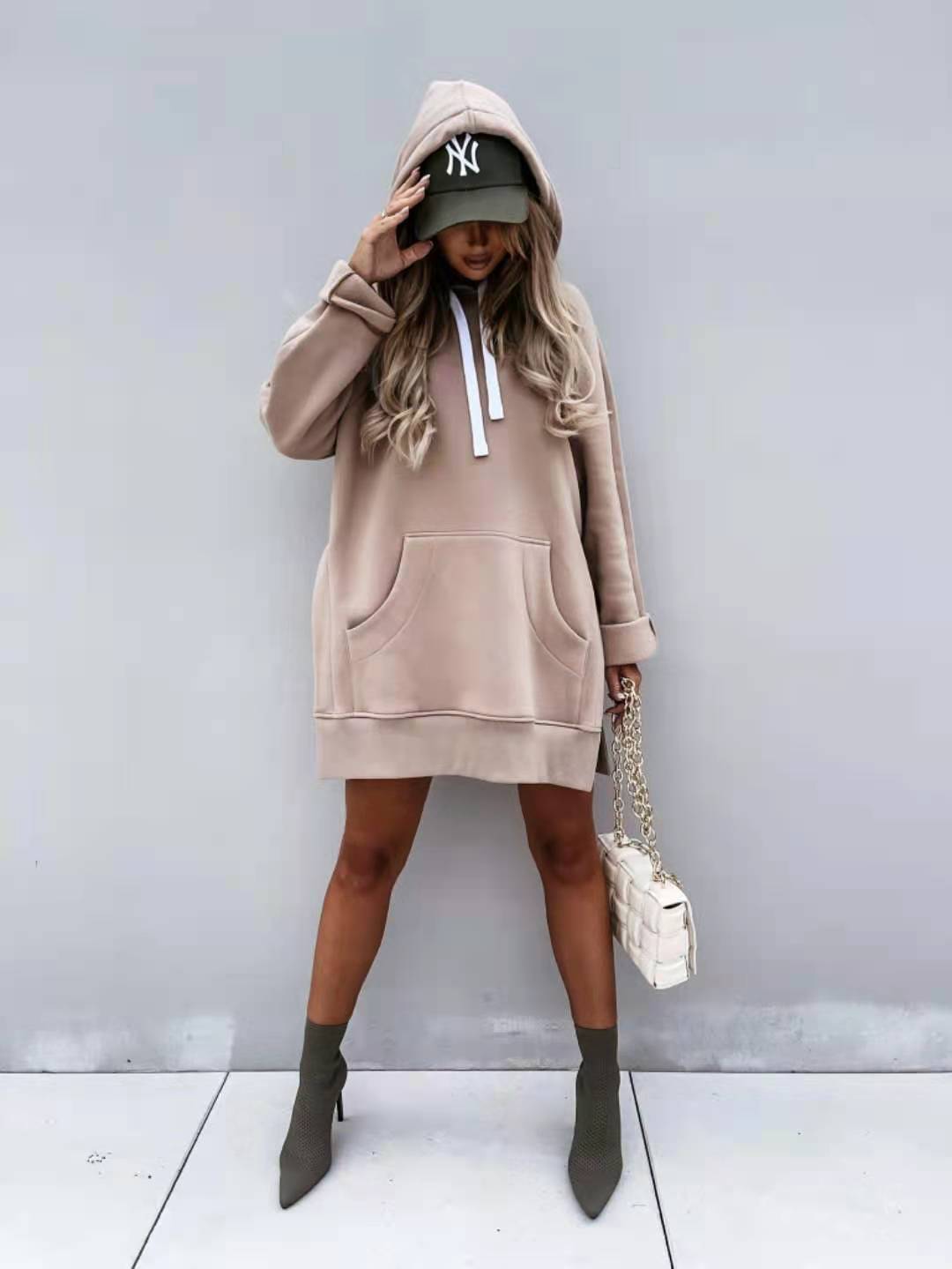 Beige oversized mid length hooded sweatshirt dress with front pockets and white drawstrings worn over a green baseball cap.