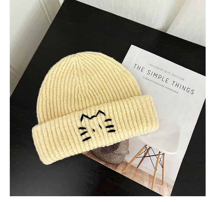 Cream-colored ribbed knit beanie hat with a black embroidered cat face on the folded brim.