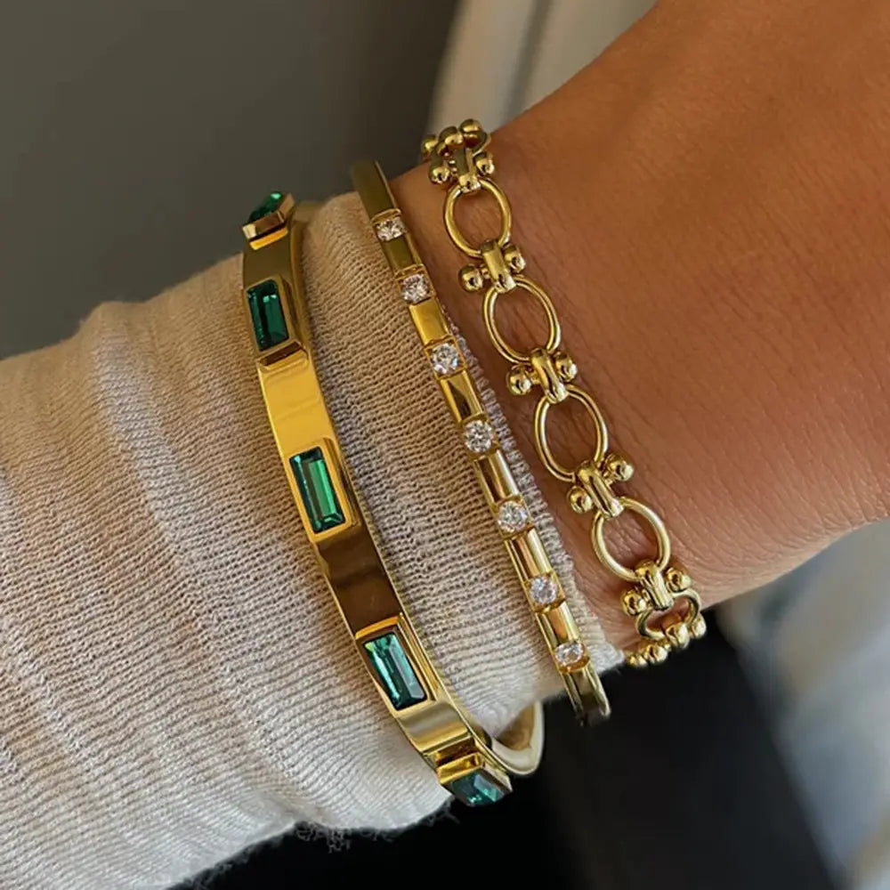 Gold toned gold plated stainless steel bangle bracelet adorned with emerald green rectangular zirconia gemstones and a delicate chain link design.