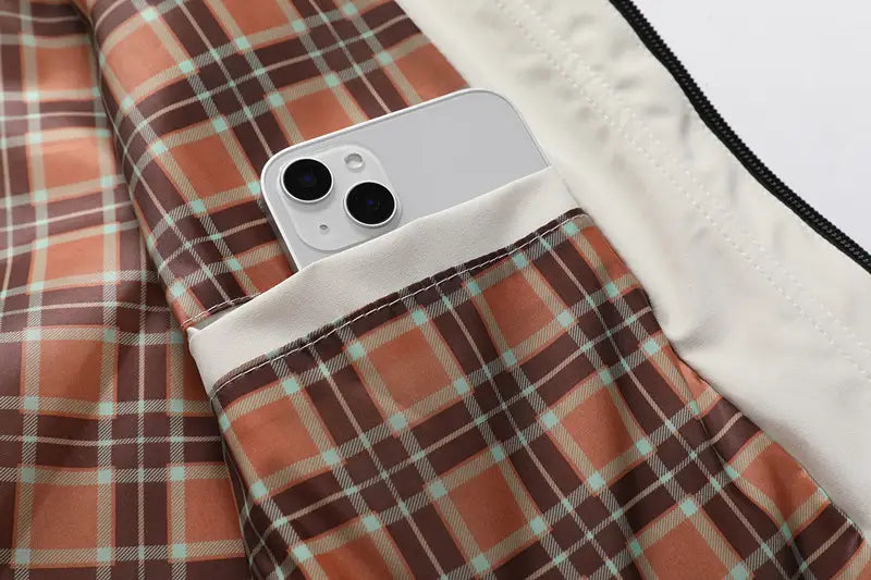 Ein silbernes iPhone 13 mit zwei Kameras, das in der karierten Tasche eines orange-braunen und hellgrünen Karojackets liegt, das unter einem Herren-Wasserabweisenden Trenchcoat mit Kapuze getragen wird