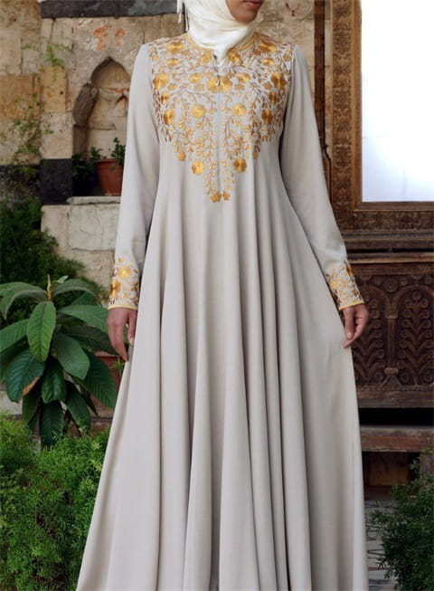 Robe abaya en coton gris clair, ample et fluide, présentant une broderie florale dorée complexe le long de l’encolure et des poignets.