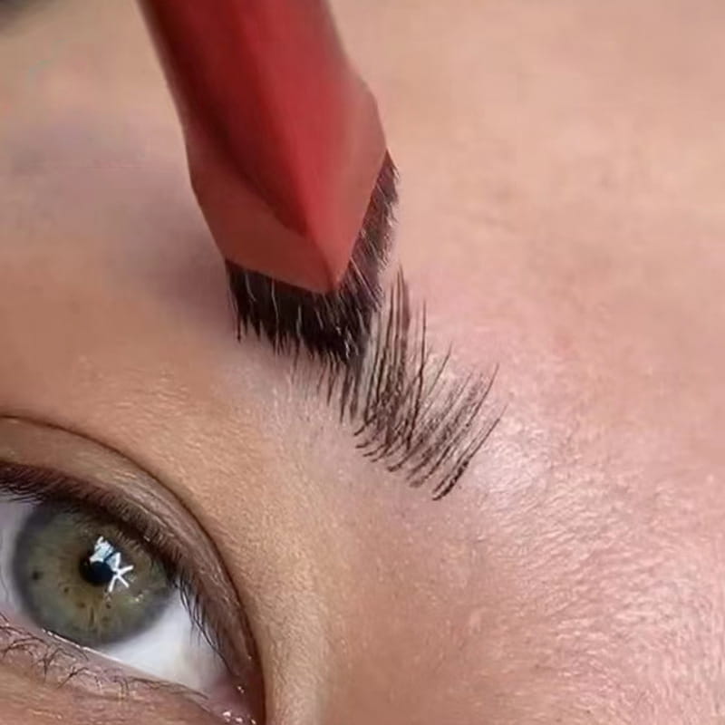 red-handled eyebrow brush with dense dark bristles being used to groom and shape brow hairs from an eyebrow brush and gel set with pink angled applicator.