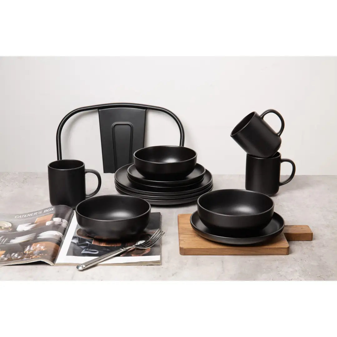 juego de vajilla de gres de 12 piezas en cerámica mate negro, con tazones, platos y tazas a juego dispuestos sobre una encimera para tres personas