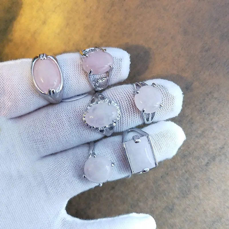 A hand wearing a white glove displays five natural gemstone rose quartz silver tone statement rings with oval round and rectangular stone shapes.