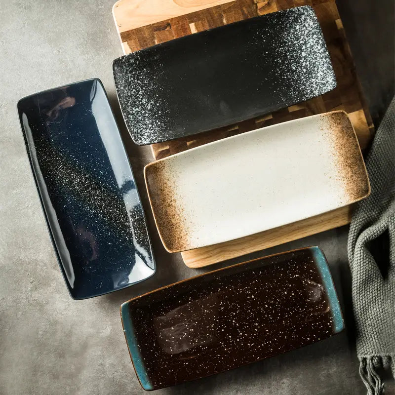 Four rectangular Japanese ceramic sushi plates in deep navy black with white speckles off-white with brown gradient and dark brown with light speckles featuring a glossy glaze finish.