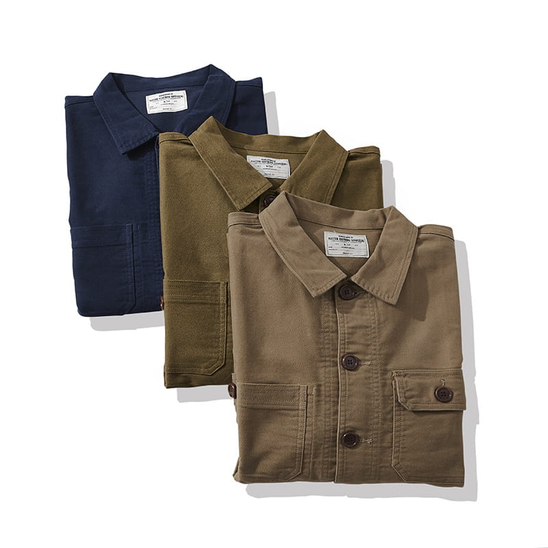 Three neatly folded mens French workwear jackets in navy olive green and tan made of cotton featuring buttoned collars and utility chest pockets.