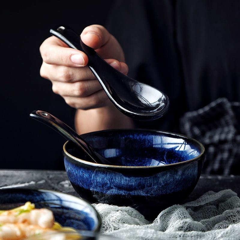 A glossy black ceramic spoon with a smooth reflective surface is held above a deep blue glazed ramen bowl ceramic 8.5 inch cat eye blue noodle bowl.