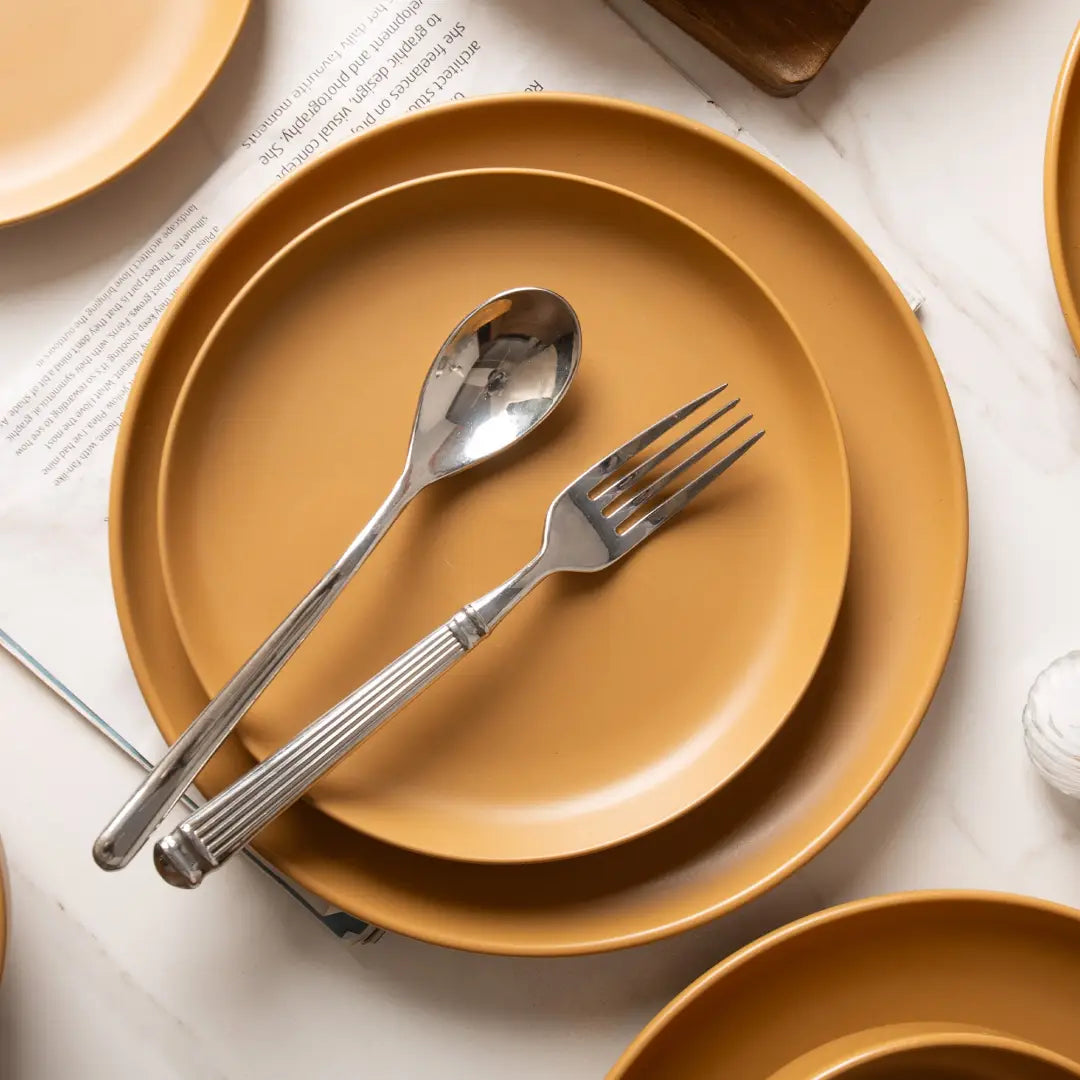 stack of mustard-yellow stoneware ceramic plates with matte glaze and silver-toned cutlery resting on top from a 12 piece dinnerware set for four