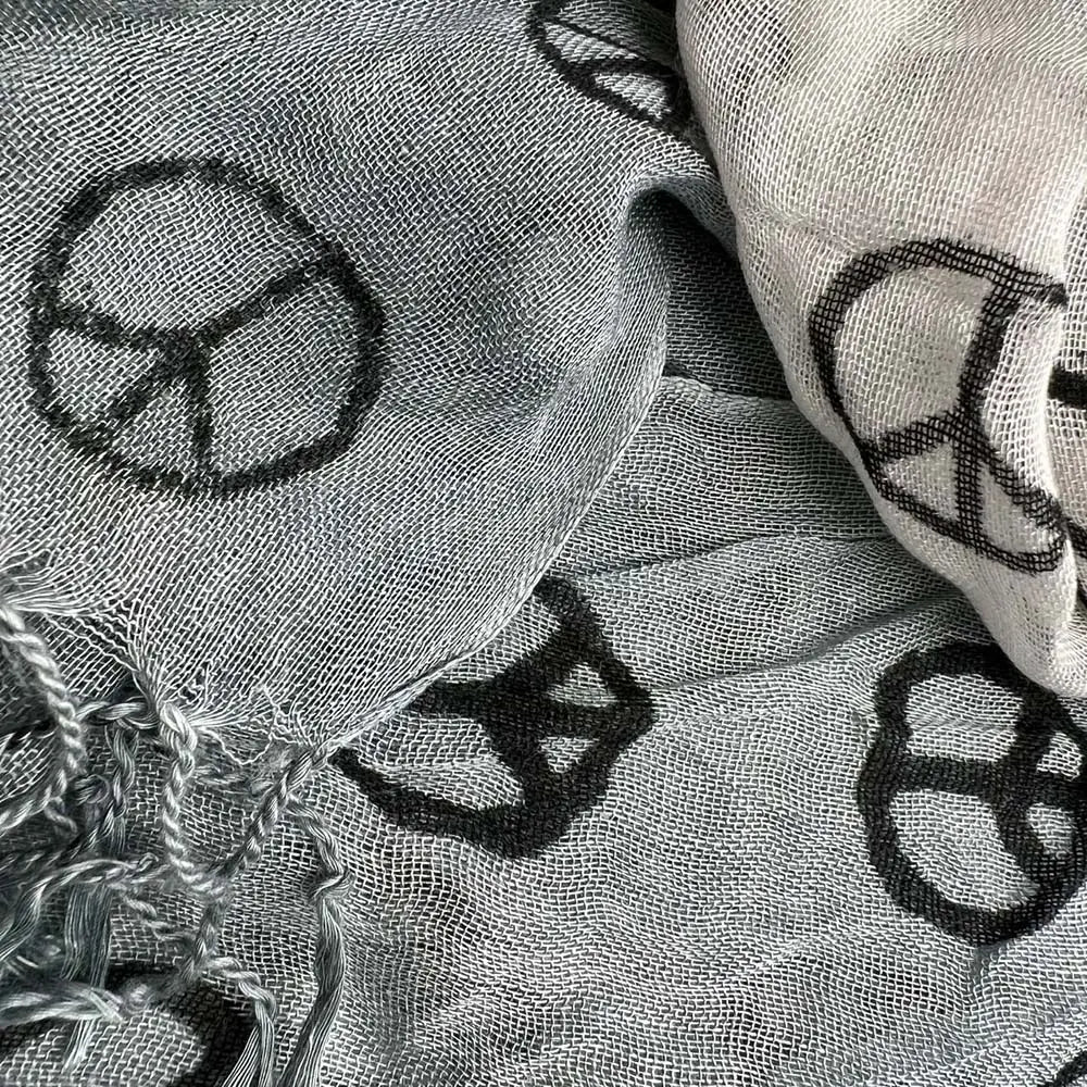 Ein hellgrauer Viskose-Schal aus locker gewebtem Stoff mit einem wiederkehrenden Muster aus schwarzen Peace-Symbolen und ausgefransten Kanten, genannt „grauer Peace-Symbol-Viskose-Schal“.