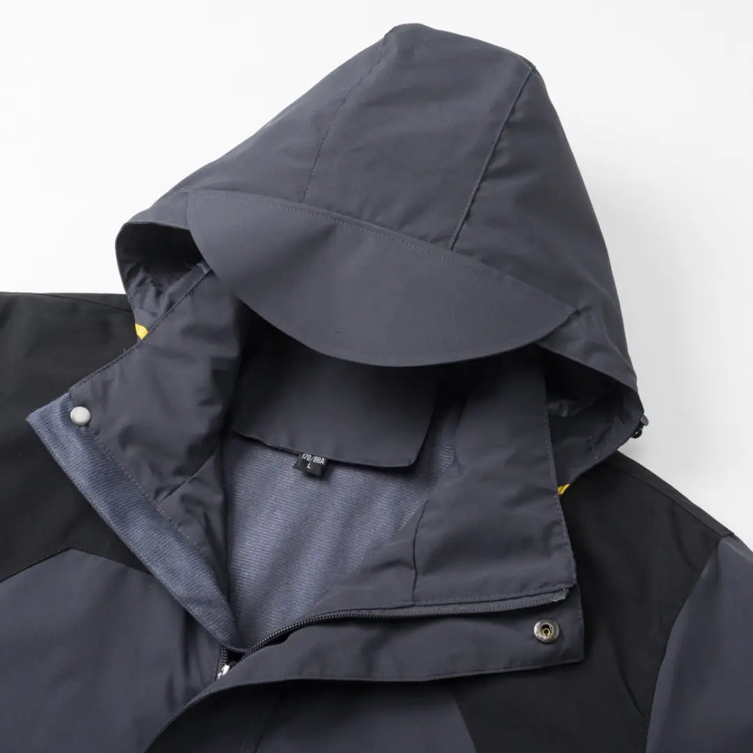 Veste de plein air imperméable à capuche gris foncé pour hommes et femmes, avec une visière structurée légèrement incurvée et des accents jaunes discrets au niveau du col.