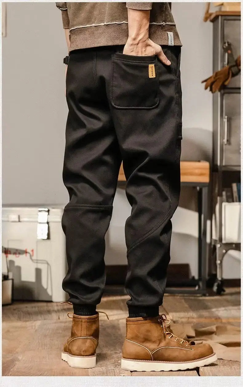 Pantalons cargo pour hommes à coupe ample, style décontracté et vêtement de travail, en denim noir, avec une texture discrète, des coutures renforcées et une poche arrière comportant un petit écusson en cuir tan, présenté depuis le côté arrière.