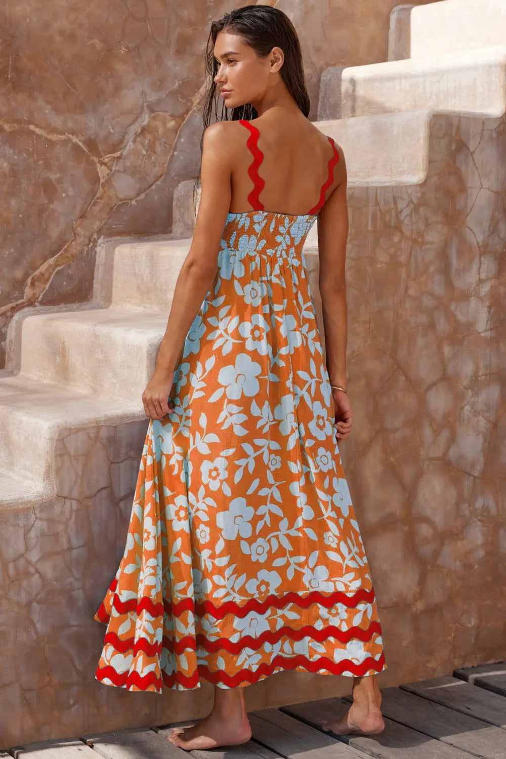 Una mujer lleva un vibrante vestido maxi de playa naranja con dobladillo ondulado floral, patrones florales celestes y ribete ondulado rojo en el contorno de las tiras, el bajo y la falda con volantes.