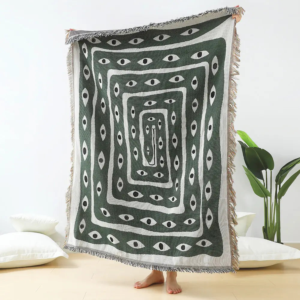 A woven cotton daisy and geometric floral throw blanket in dark green featuring a bold white geometric pattern of concentric squares filled with stylized eyes and fringed edges.