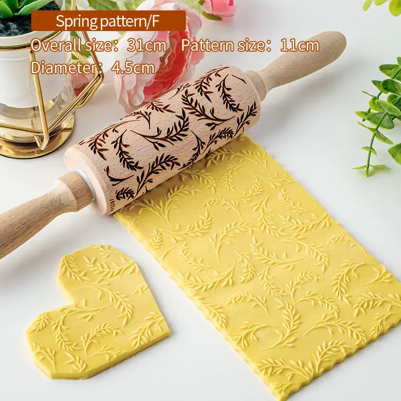 Un rouleau à pâtisserie en bois de hêtre en relief floral présentant un motif printanier sombre brun et des poignées en bois clair, utilisé pour imprimer des motifs feuillus sur de la pâte jaune.