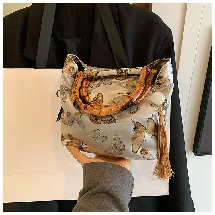 A light gray bamboo handbag with tassel detail and butterfly print canvas featuring bamboo handle accents.