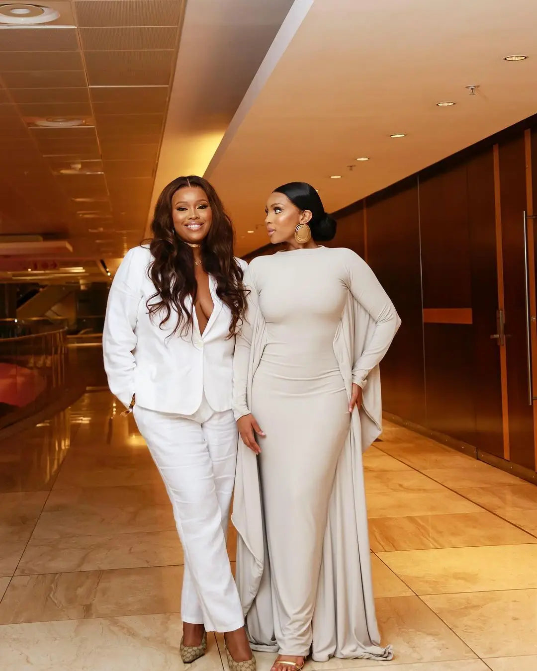 The women are wearing elegant asymmetrical satin batwing sleeve maxi dresses in white and light gray formal attire.