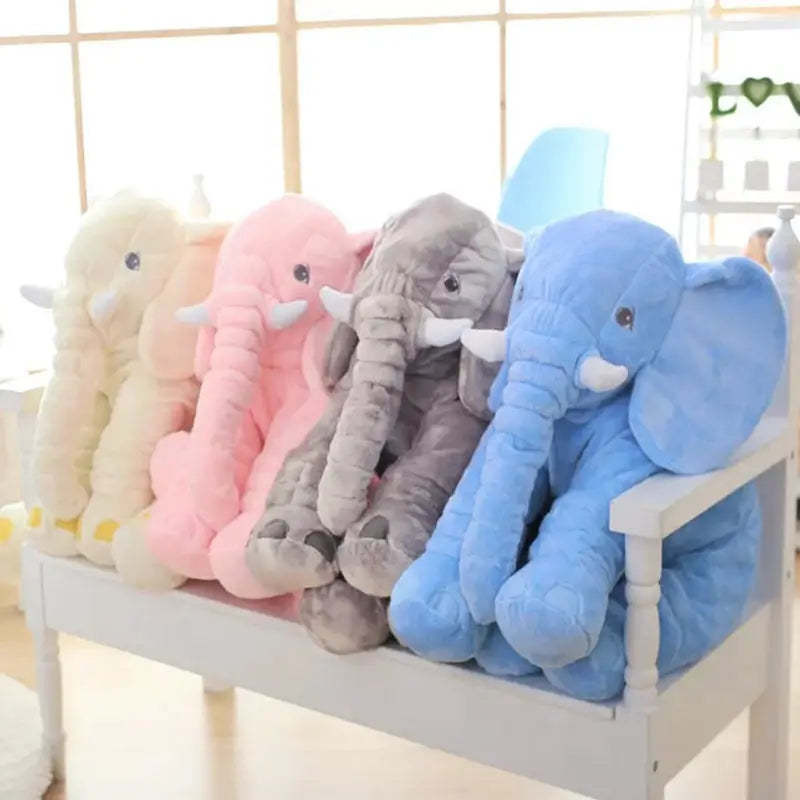 Coussins d’animaux en peluche représentant des éléphants, en peluche douce, dans des tons pastel crème, rose, gris et bleu, disposés sur un banc blanc pour les enfants.