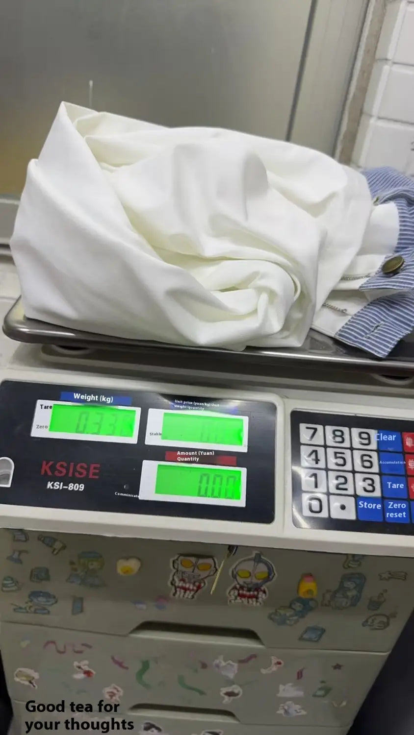 Chemise en coton blanche légèrement froissée, avec poignets rayés bleu et blanc, posée sur une balance numérique à côté d’un jean à taille avec bande blanche et jambes évasées.