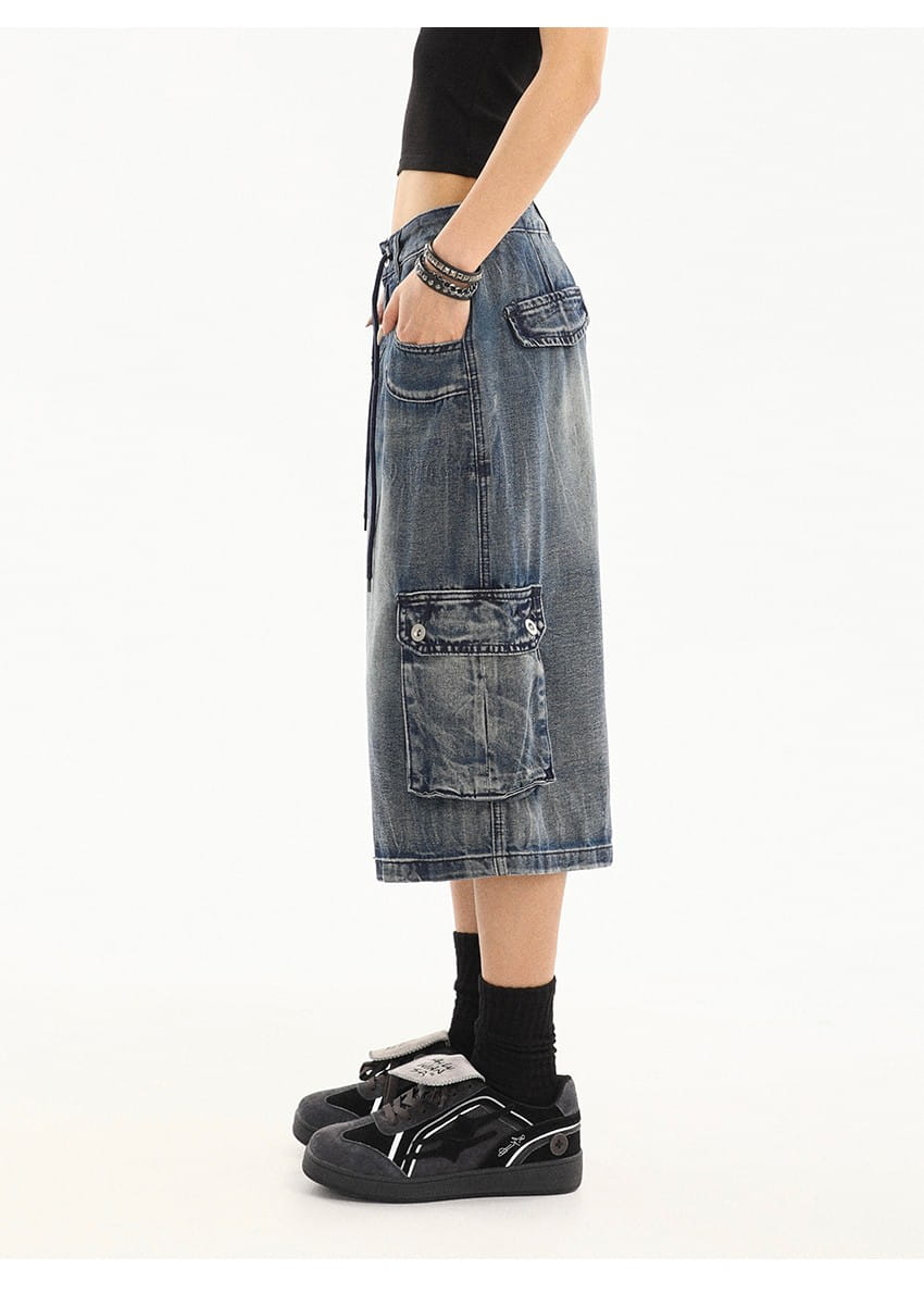 Falda de mezclilla azul de largo a la mitad del muslo, con lavado envejecido, múltiples bolsillos tipo parche y cinturón con cordón, diseñada como pantalones cargo urbanos de estilo con pierna recta.