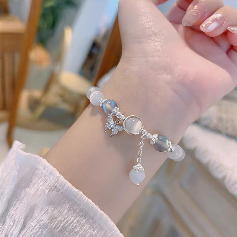 A delicate silver-toned crystal butterfly cat’s eye bead bracelet featuring translucent light blue and white beads, accented with a sparkling butterfly charm and a dangling pearl-like drop.