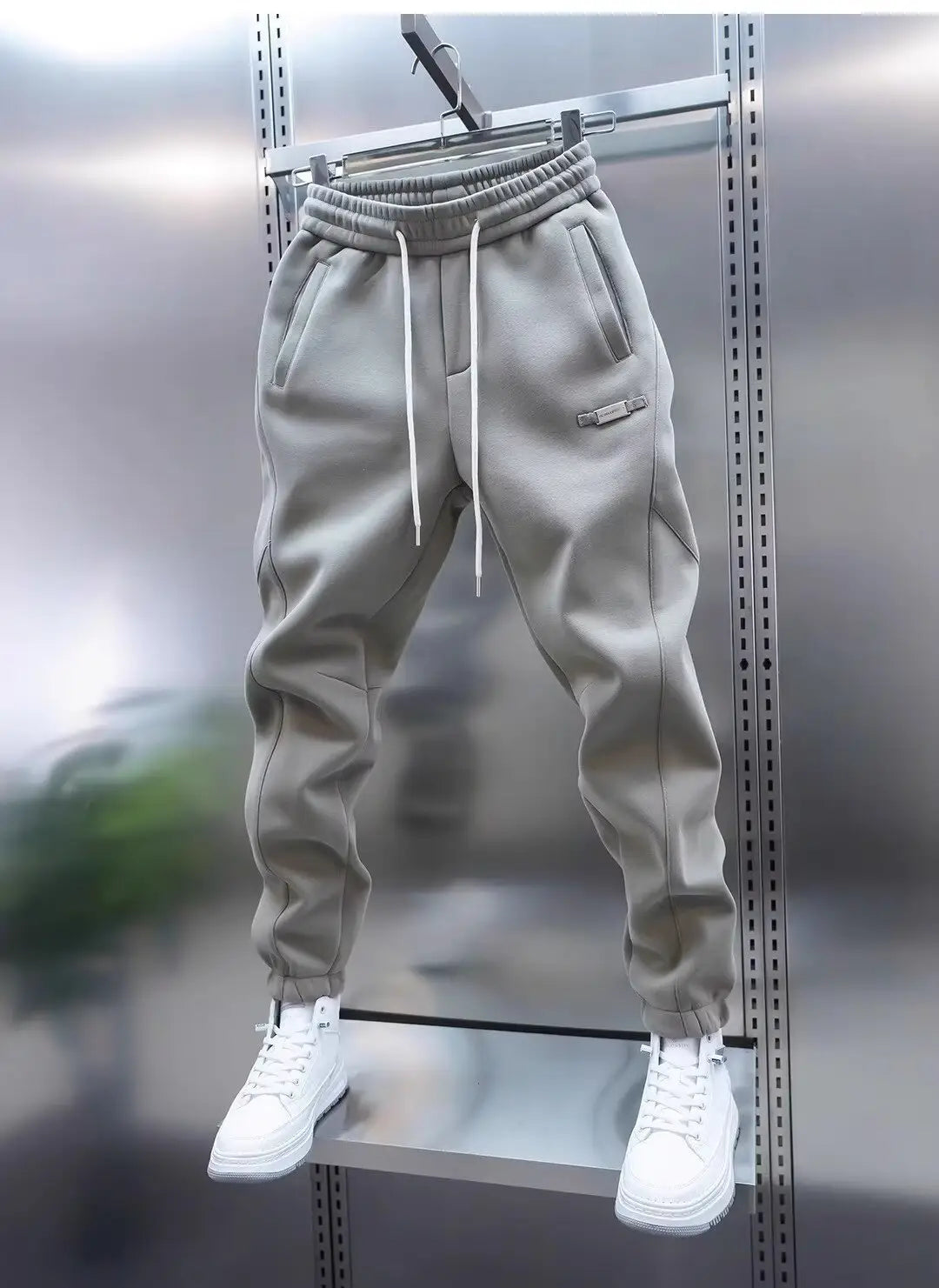 Mens casual solid color sweatpants in gray with white drawstrings and a subtle logo patch on the thigh hanging neatly against a metallic backdrop.