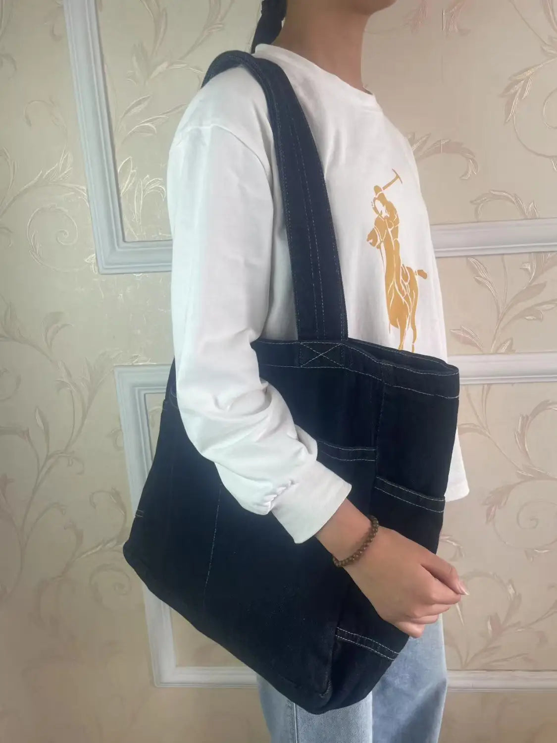 Black denim tote bag with contrasting white stitching and a sturdy shoulder strap from Alma Latina featuring large capacity and durable material.