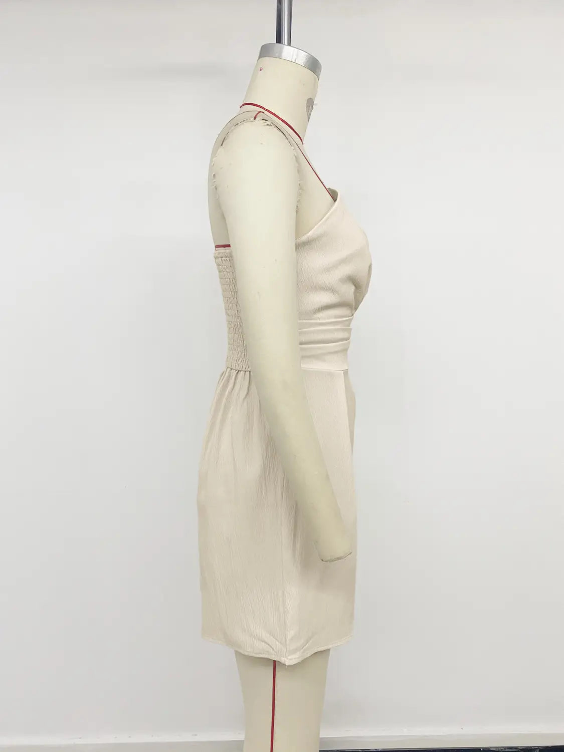 Beige sleeveless dress with subtle smocking at the bodice and thin red piping along the neckline and side seam resembling a cream strapless pleated jumpsuit.