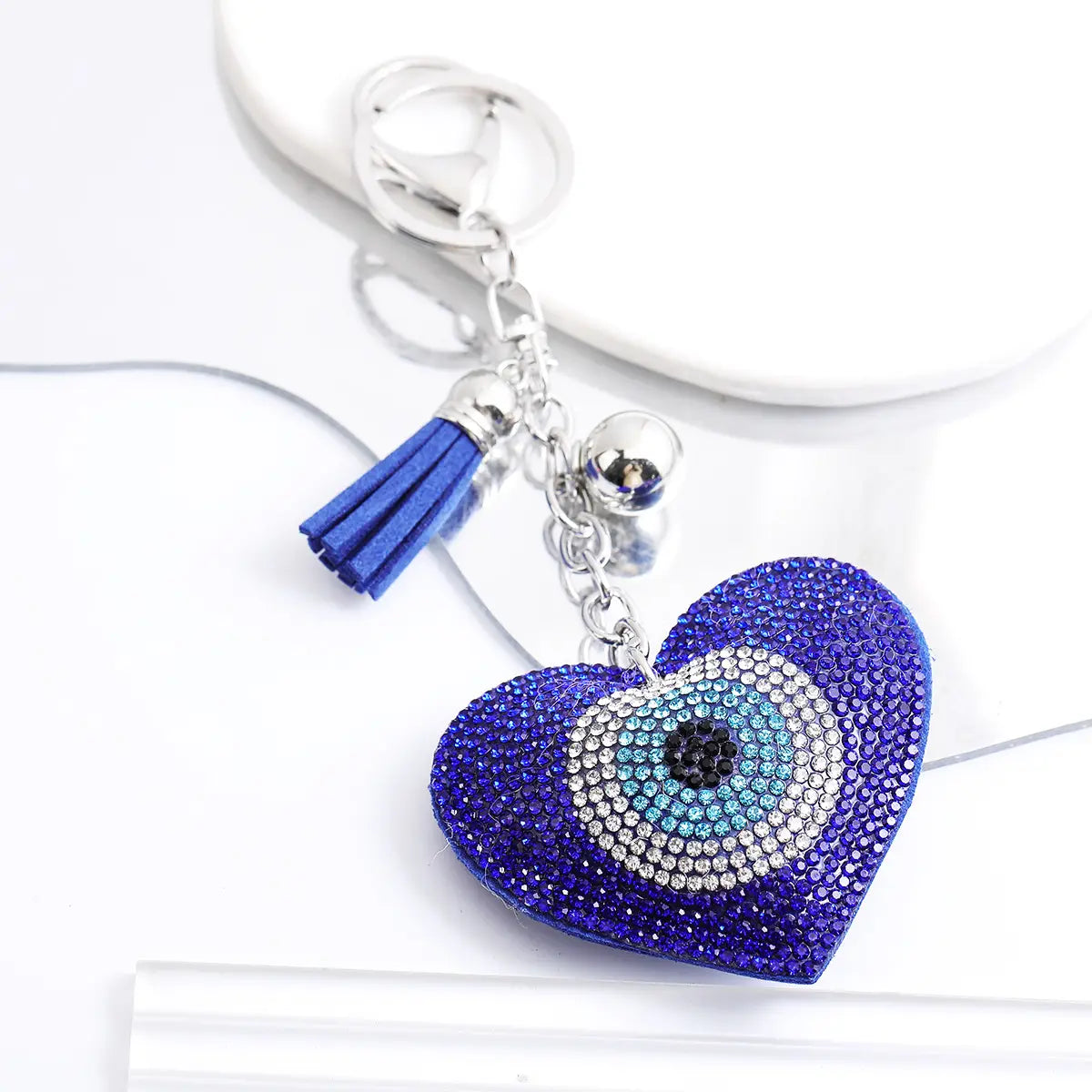 A vibrant blue heart-shaped leather keychain adorned with sparkling rhinestones and a central evil eye design featuring a silver chain, tassel, and bell.