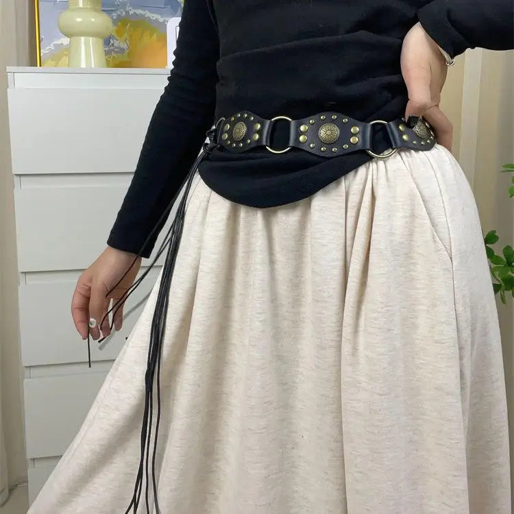Schwarzer Gothic-Punk-Lederharness-Gurt mit messingfarbenen, kreisförmigen Nieten, Metallringen und baumelnden schwarzen Quasten.