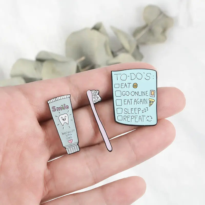 A light blue enamel pin shaped like a to-do list with checkboxes and doodles of food items in the toothpaste smile enamel pin cartoon tooth brooch design.