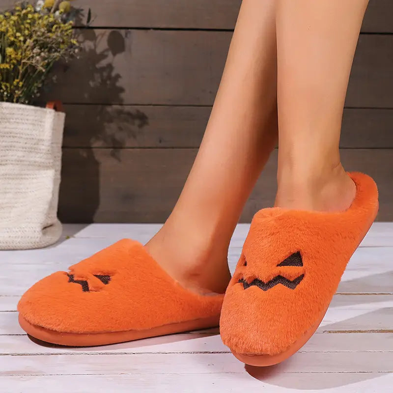 Orange plush pumpkin-themed slip-on indoor slippers with black embroidered facial features for Halloween.