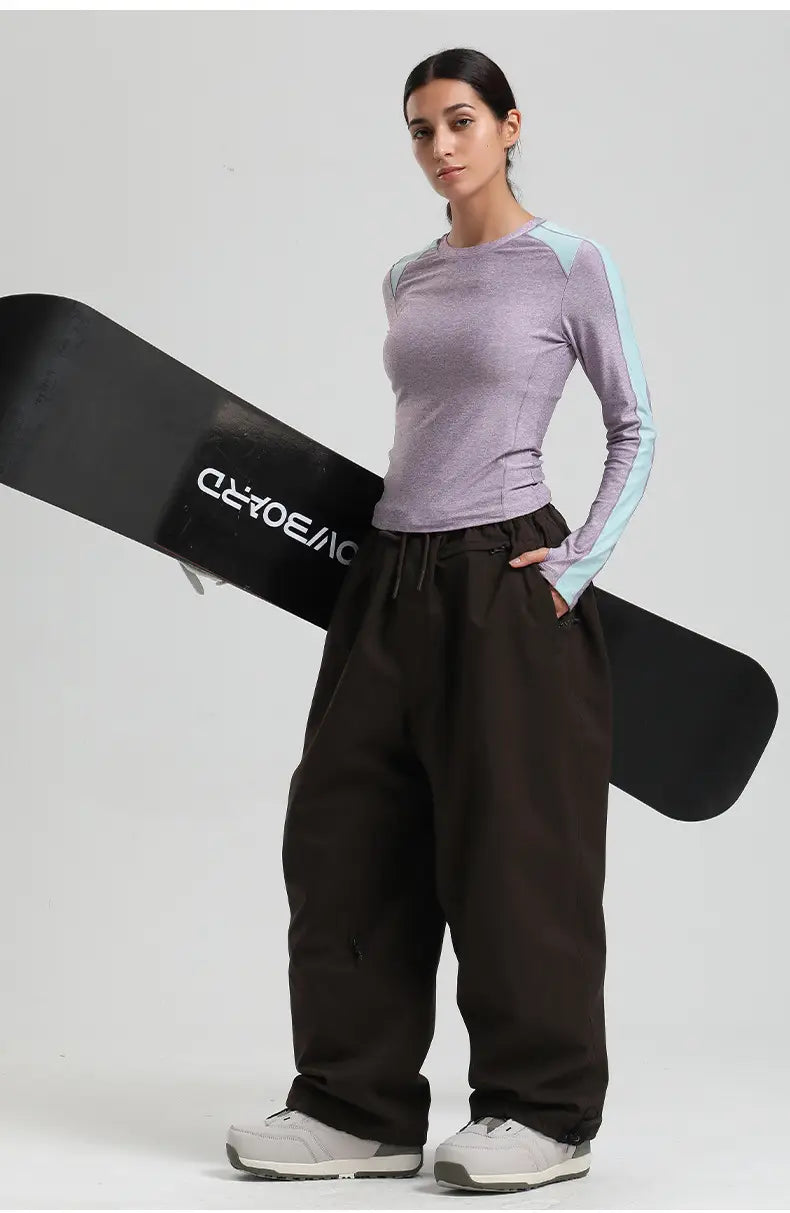 The image shows a black snowboard with white crossboard lettering next to waterproof windproof loose black snow pants designed for work and outdoor use.