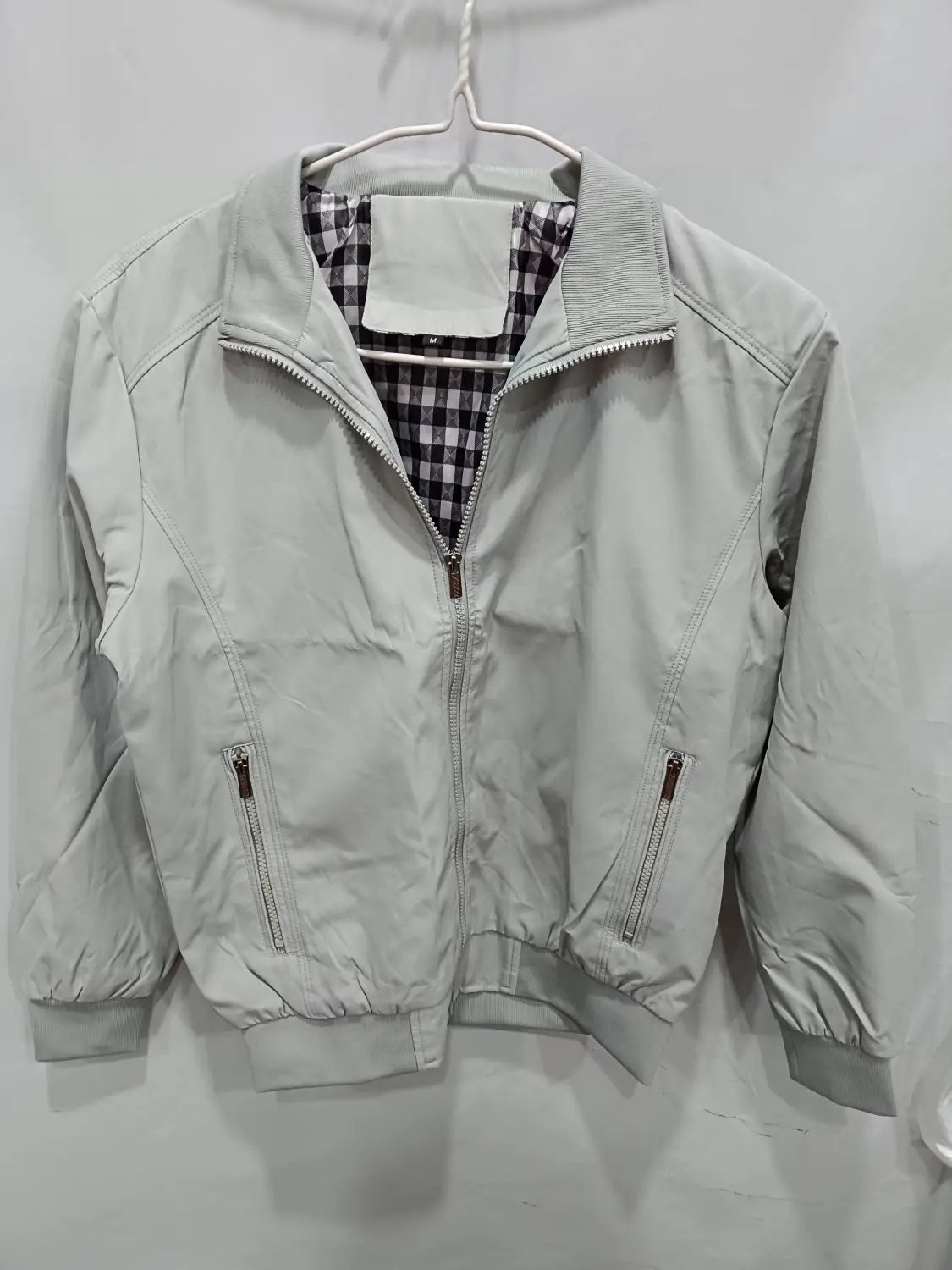Veste bombardier légère pour homme en gris clair, avec doublure à carreaux noir et blanc et deux poches avant zippées.