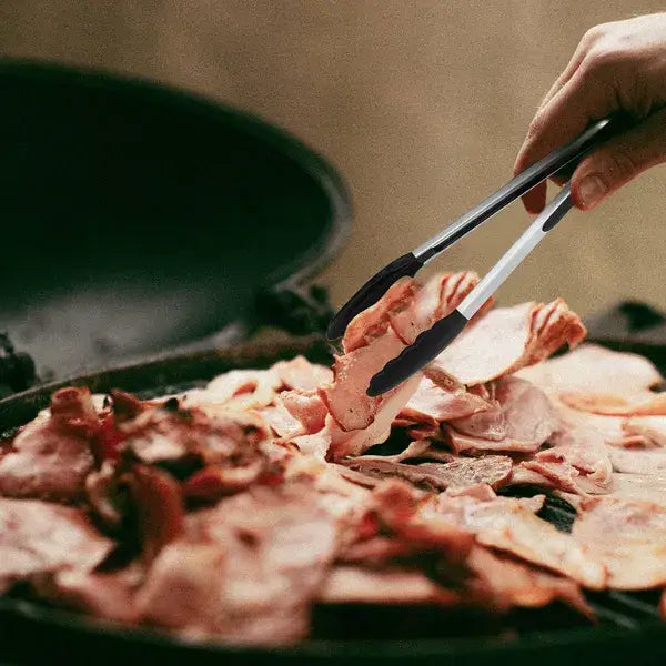 Black and silver stainless steel kitchen tongs with silicone tips holding slices of raw pork belly over a grill.