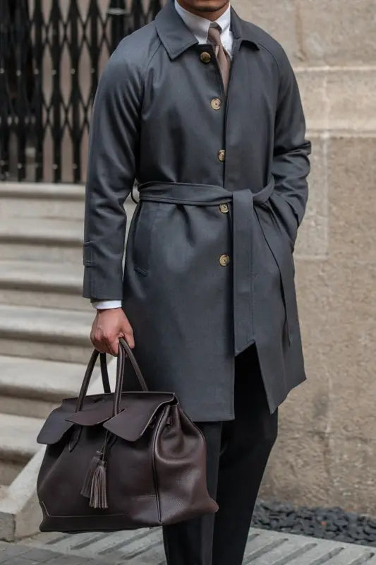Dark brown leather satchel with flap closure and tassel detail complementing a wool winter trench coat single-breasted dark gray.